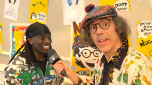 Kai Cenat smiles while being interviewed by Nardwuar, who holds a microphone and wears his signature plaid hat, in a vibrant setting with colorful posters in the background.