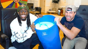 Kai Cenat and Mark Rober smiling and holding a large blue bucket filled with a mysterious liquid during a fun science experiment.